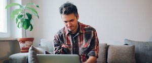 Young man working on a laptop, sitting on his sofa, having a cup of coffee.