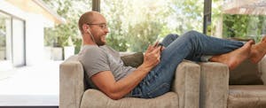 Man with earbuds in his ears sitting across an armchair smiles while he looks at a tablet screen.