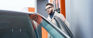 Man with glasses and a beard opening his car door at a gas station and contemplating the costs of becoming a Lyft or Uber driver.