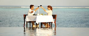 Couple sitting at a table in the surf at a beach, toasting before enjoying a meal together