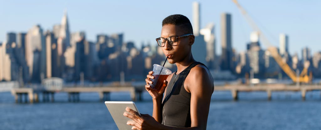 Woman on a boardwalk, sipping a cold drink, and using the best ways to send money transfers online on her tablet