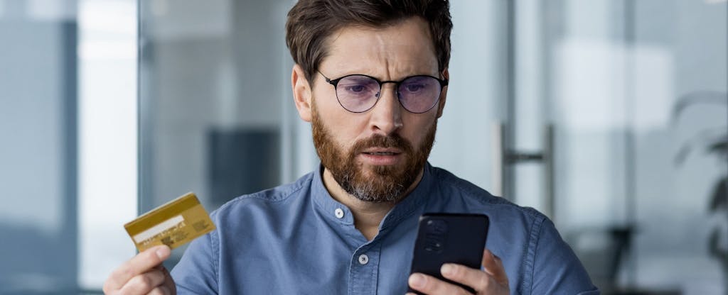 Upset young male businessman appears worried while holding credit card and mobile phone, sitting in office at desk.