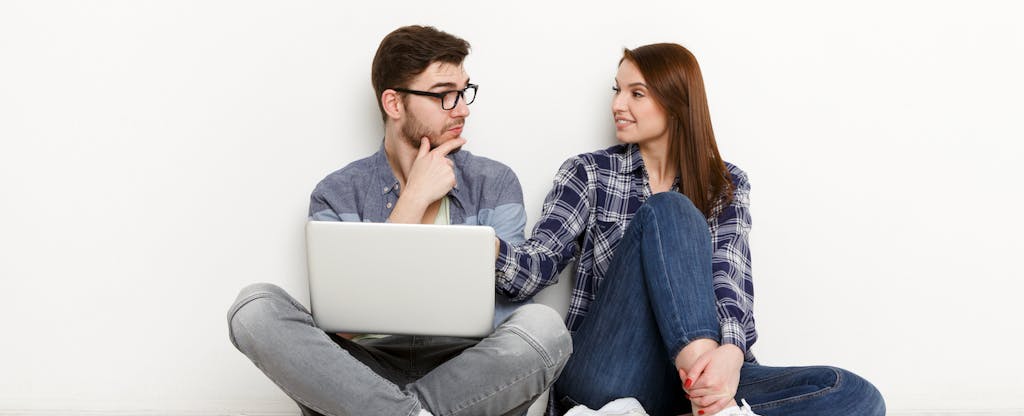 Young couple leaning against a white backdrop with a laptop while discussing whether they should refinance their mortgage