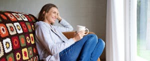 Woman sitting on a cozy couch, drinking coffee, and thinking about how to pay off debt by cutting her expenses