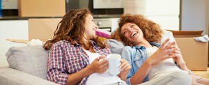 Two women laugh on the sofa in their new house after getting their debt-to-income ratio increase.