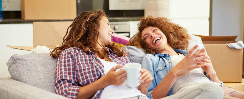 Two women laugh on the sofa in their new house after getting their debt-to-income ratio increase.