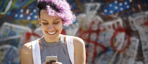 Smiling young woman with purple hair using her smart phone against graffiti wall