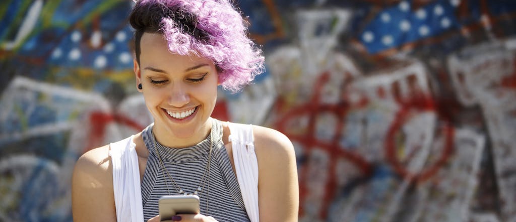 Smiling young woman with purple hair using her smart phone against graffiti wall