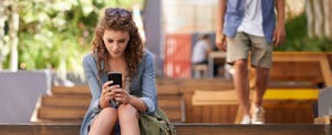 Young woman sitting on steps in a marketplace, looking purposefully at the screen of her smartphone in her hands