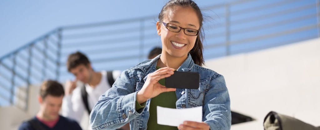 Happy young Asian-American woman using her smartphone to deposit her income by snapping a picture of her paycheck.