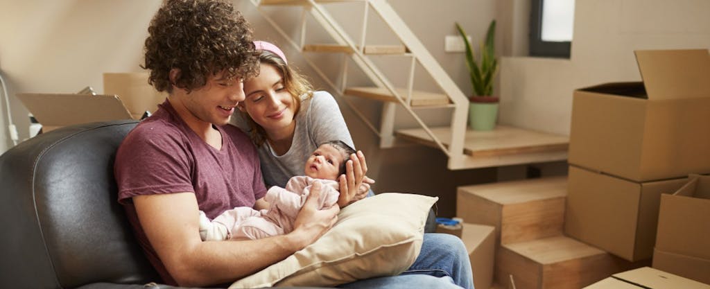 Young man and his wife, holding their newborn and relaxing in the living room of their new home amid moving boxes.