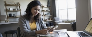 Young woman at dining room table, counting how much cash she will save by using free tax filing software instead of paying to file taxes.