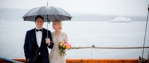 A smiling bride and groom stand outside on a deck under an umbrella on a rainy day.