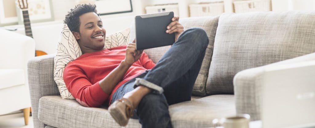Young African-American man, lying on sofa, e-filing his taxes on a tablet.