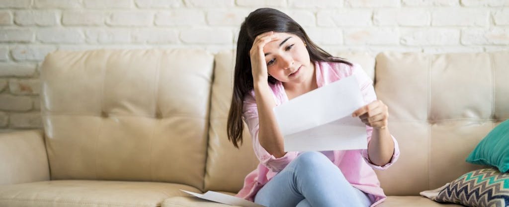 Young Hispanic woman sitting on couch, looking worried, as she reads a letter informing her her tax refund will be offset.