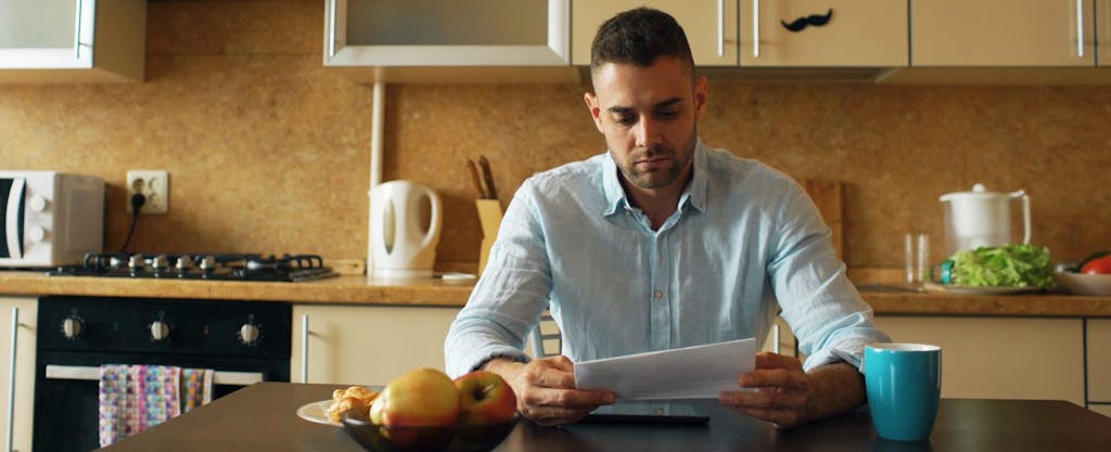 Worried young man sitting at his kitchen table and reading a letter from the IRS.