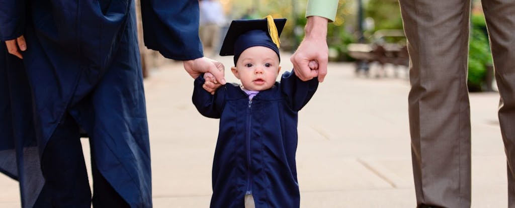 Close up of cute baby in blue graduation cap and gown, holding hands with mom and dad.