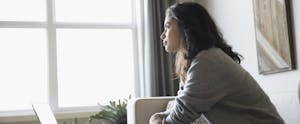 Pensive young woman at laptop looking through window in living room