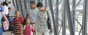 Family of four walking together in airport, mother and daughter in front, holding hands