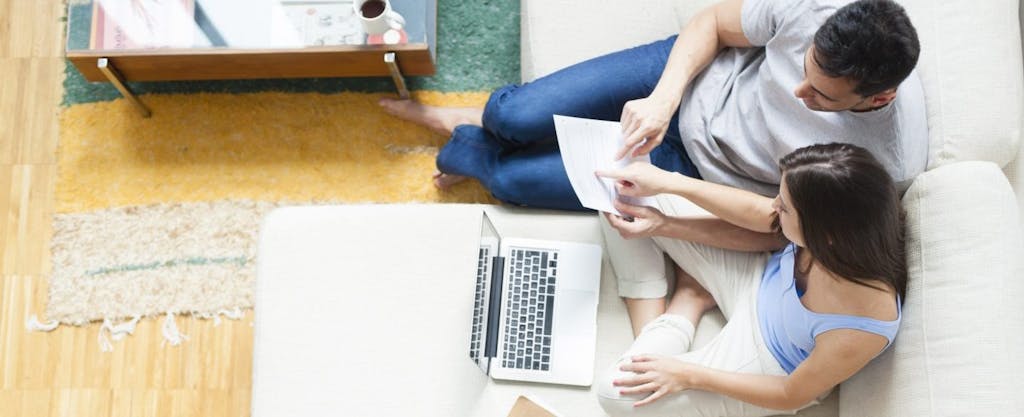 Overhead view of young couple, sitting on couch, and working on a lap top to estimate their income taxes.