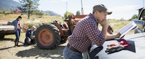 Farmer leaning on the hood of his truck, tractor and field in the background, reviewing tax forms.