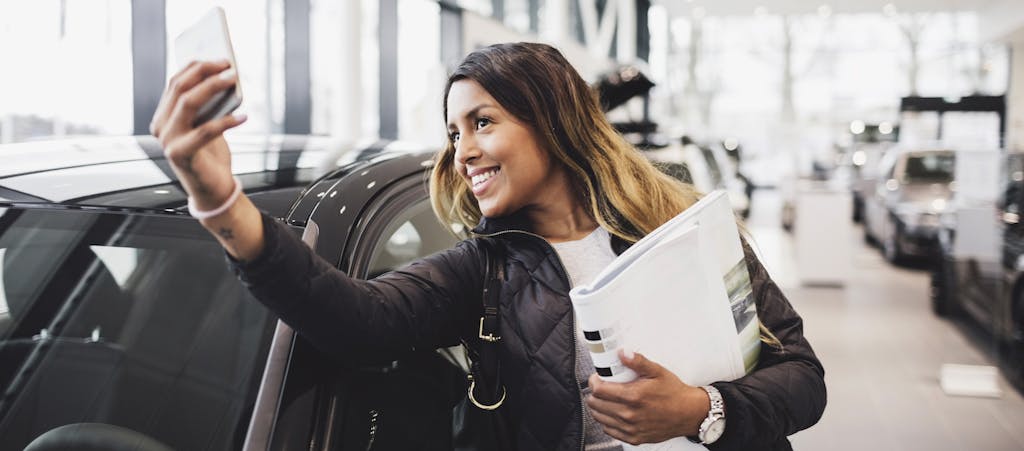 Smiling woman taking selfie with new car in showroom