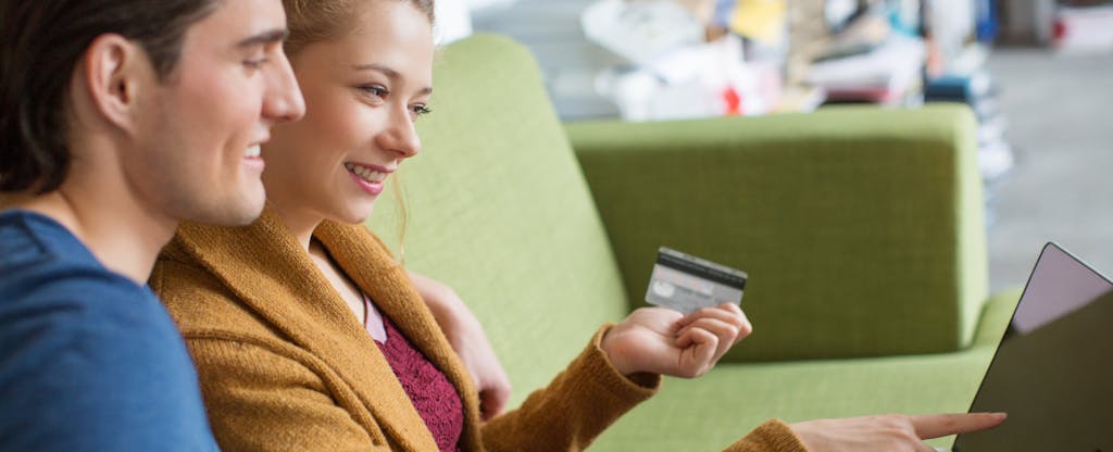 Happy young couple sitting together on sofa shopping online together using a laptop and credit card