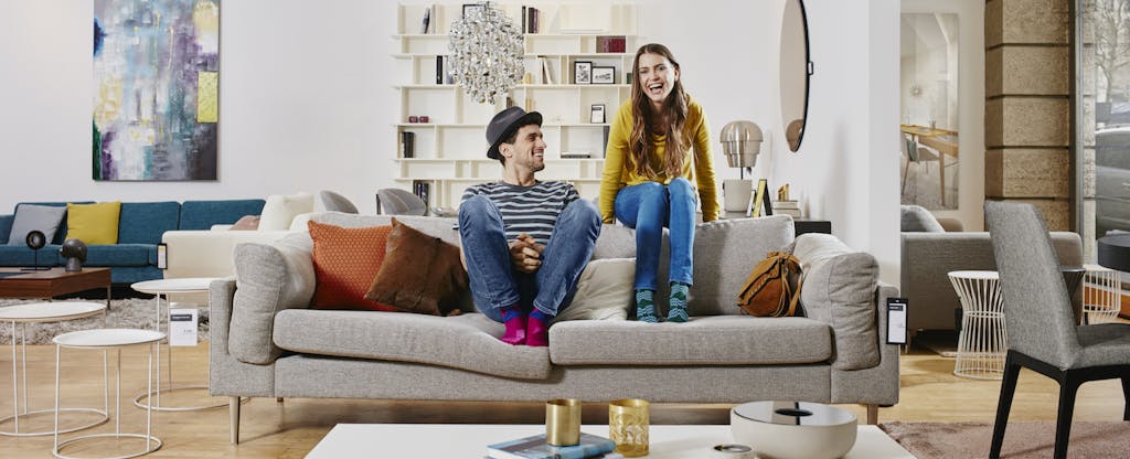Couple in modern furniture store sitting on couch, laughing and wondering about the difference between their FICO score and credit score.