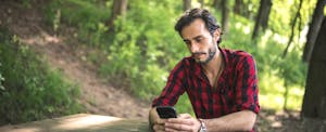Young man in red and black plaid shirt, sitting at a park bench, looking sadly at his phone.