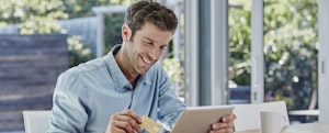 A blond man holds a credit card and a tablet and smiles about knowing how long a balance transfer will take.