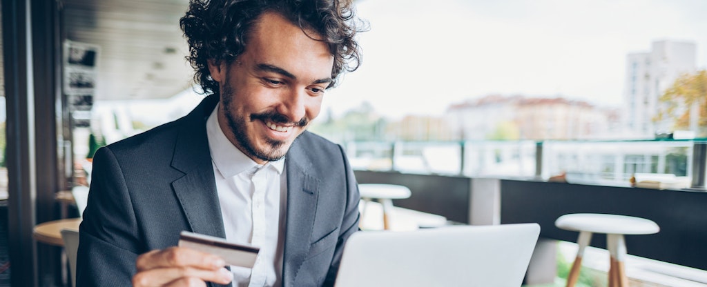 Smiling man sitting in cafe, holding a credit card and typing on a laptop.