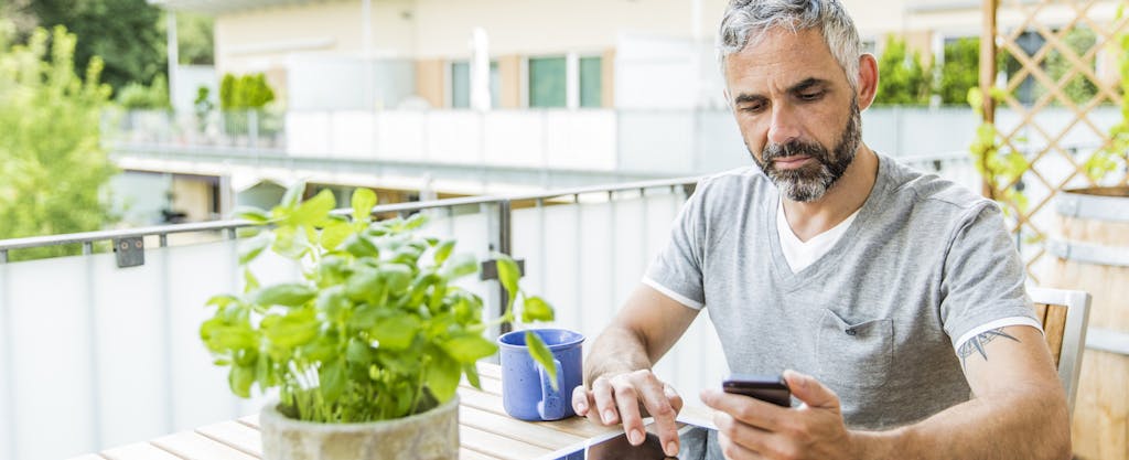 Portrait of man sitting on his balcony using smartphone and digital tablet
