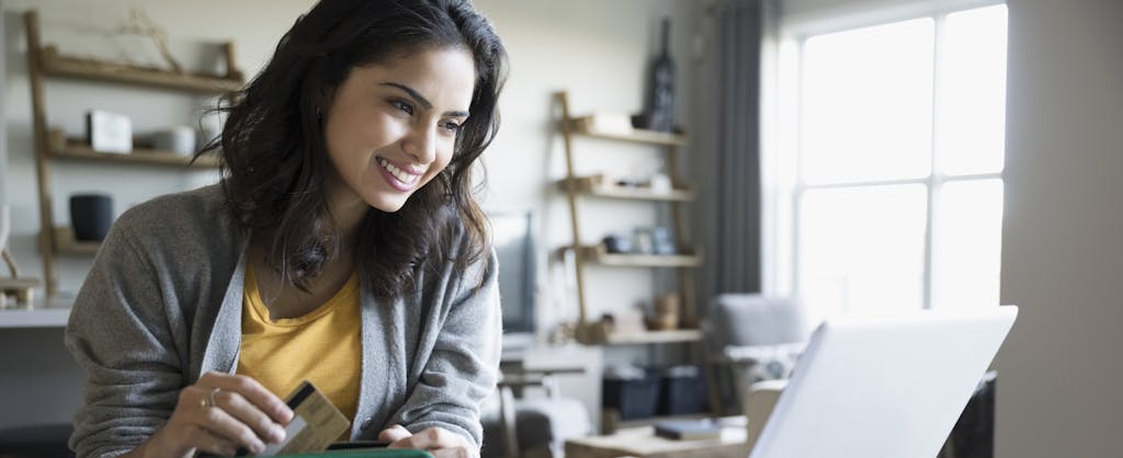 Smiling young woman with credit card and laptop learning about balance transfers in dining room