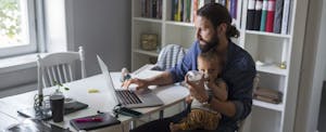 Millennial father with man bun holding baby on his lap, looking at laptop and trying to decide if you should file as head of household on his 2018 tax return.