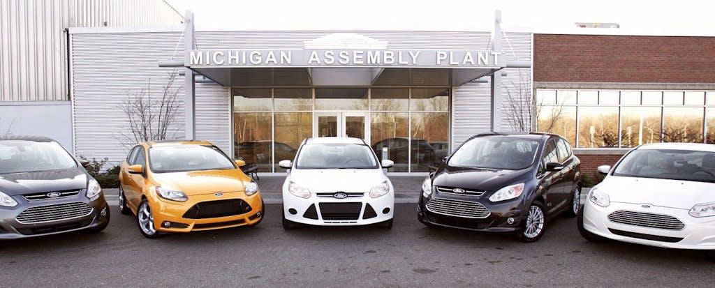 A row of new cars sits in front of a Michigan auto assembly plant.