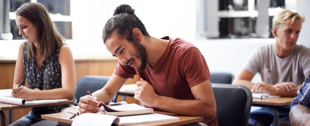 Young college student with a beard and man-bun, working at classroom desk and wondering what education credits he might be eligible for.