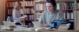 Young male college student with dreads sitting at a library table, calculating what school-related costs might be qualified education expenses for tax breaks.