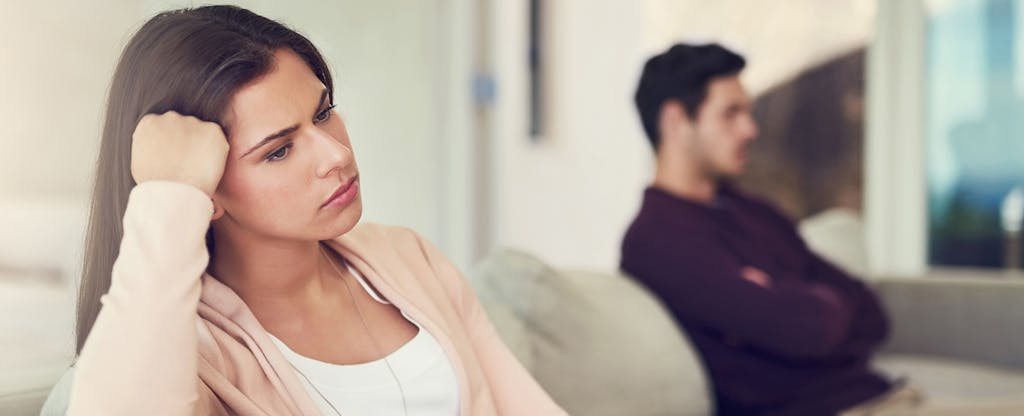 Pensive young couple sitting on couch, wife in the foreground wondering if injured spouse relief can help ensure she doesn’t lose all her tax refund due to her husband’s past-due tax debt.
