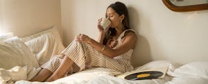 A young woman engages in JOMO while drinking coffee from cup in bed