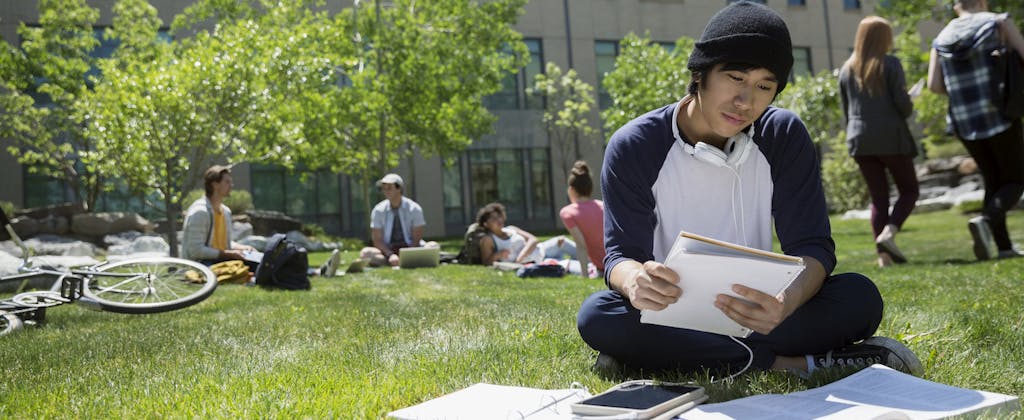 Asian-American college student, sitting on campus lawn, reviewing student loan paperwork and wondering how to get a copy of a tax return for his application.