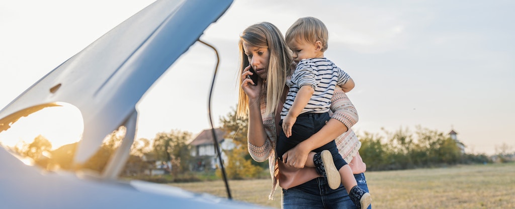 Young woman standing by broken down car and waiting for assistance while holding her baby boy and talking on cell phone