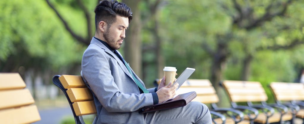 A young businessman is drinking coffee and working on a digital tablet in a public park.