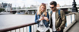 A young couple with camera standing by the river in a city, using smartphone