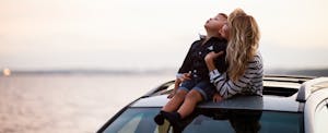 Woman standing in the sunroof of her car, holding her child, who sits on the roof of the car