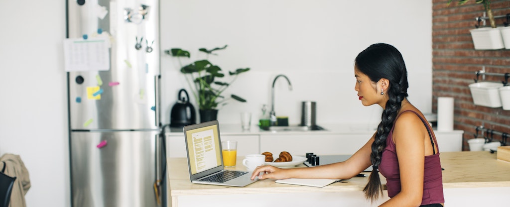 Woman sitting at her kitchen counter, eating breakfast and reading on her laptop