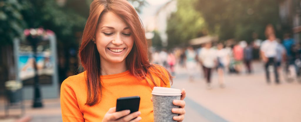Young woman walking outside, holding coffee and texting