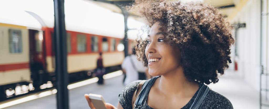 Woman standing at a train station and smiling as she uses her phone