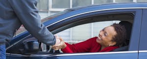 Woman sitting in the driver's seat of her car, shaking hands with a man standing outside of the car