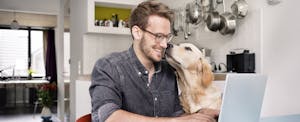 Man sitting at his kitchen table, working on his laptop, while his dog licks his face