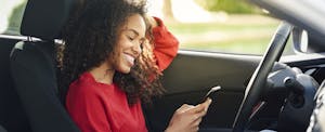 Smiling young woman sitting in her car and looking at her cell phone
